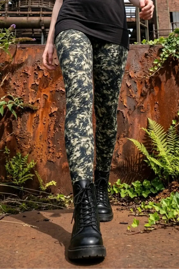Person trägt grün gemusterte Leggings und schwarze Stiefel vor rostiger Metallwand, Outdoor-Setting.