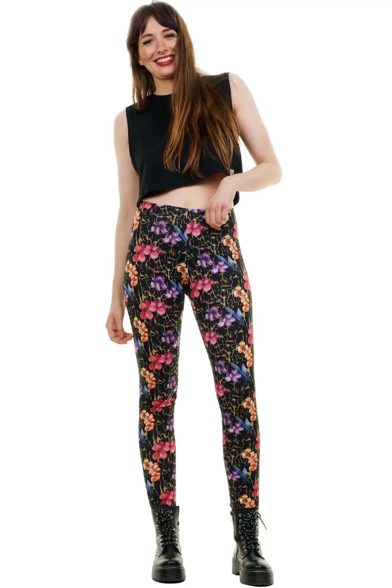 Ganzkörperansicht eines Models in blickdichten, schwarzen Baumwoll-Leggings mit buntem Blumenmuster. Kombiniert mit einem schwarzen ärmellosen Crop-Top und klobigen Schnürboots vor weißem Hintergrund. Model fasst sich lächelnd in die Haare und blickt zur Seite